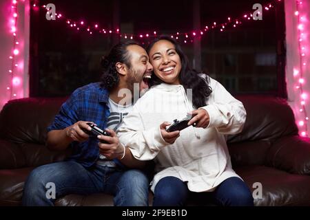 Eccitata coppia etnica in abbigliamento casual con giochi di gassy pads videogioco insieme mentre sei seduto sul divano in pelle a casa Foto Stock