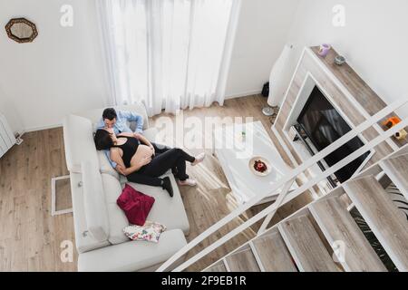 Dall'alto della donna incinta seduta sul divano con l'uomo e baciandosi mentre godendo il fine settimana insieme a casa Foto Stock
