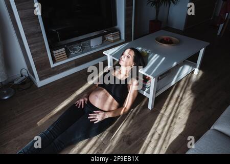 Alto angolo di tranquilla donna incinta appoggiato sul tavolo e. seduto sul pavimento a casa mentre si gode la luce del sole con chiuso occhi Foto Stock