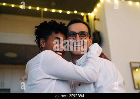 Vista laterale basso angolo di soddisfare multirazziale coppia abbracciando mentre in piedi in camera con ghirlata illuminata Foto Stock