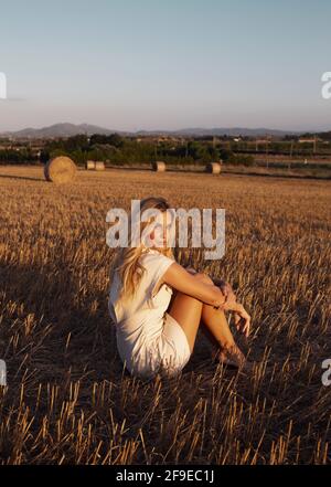 Donna tranquilla in abito elegante seduta su campo asciutto dentro zona rurale e guardando la macchina fotografica Foto Stock