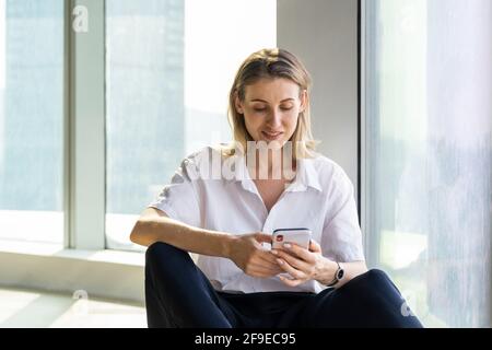 Delight giovane donna seduta in un ufficio vuoto con grande finestra di navigazione sul telefono cellulare Foto Stock