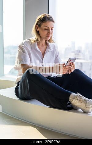 Solitaria giovane donna senza emozioni seduta in un ufficio vuoto con grande finestra di navigazione sul telefono cellulare Foto Stock