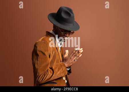 Vista laterale di un giovane maschio afroamericano in abbigliamento alla moda e cappello guardando via su sfondo marrone Foto Stock