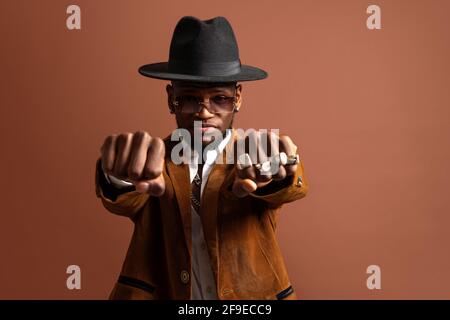 Giovane etnico maschio in cappello e abbigliamento elegante guardando fotocamera su sfondo marrone Foto Stock