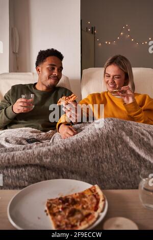 Coppia multietnica seduta in poltrone e mangiare deliziosa pizza mentre goditi il weekend insieme a casa tua Foto Stock
