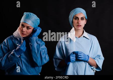 Giovani donne colleghe in uniformi mediche che togliano le maschere facciali mentre si è in piedi su sfondo nero in clinica Foto Stock