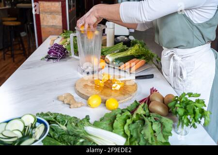 Vista laterale del prodotto femmina non riconoscibile che mette fette arancioni fresche nel recipiente del frullatore a tavola con verdure assortite in cucina Foto Stock