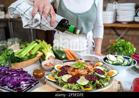 Raccolto femmina irriconoscibile versando olio d'oliva da bottiglia su yummy insalata vegetariana con verdure assortite e semi di sesamo a casa Foto Stock