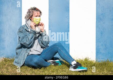 Una donna caucasica che indossa una maschera facciale e si siede sul erba contro una parete a strisce blu e bianche Foto Stock