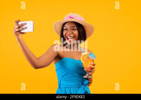 Donna nera felice con cocktail estivo yummy che prende selfie sopra smartphone su sfondo arancione dello studio Foto Stock