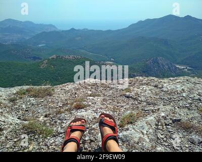 Piedi da donna in scarpe da trekking, illuminati dal sole su una scogliera sullo sfondo di montagne e mare blu Foto Stock