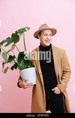 Uomo di mezza età sicuro in corto cappotto beige, pentola di tenuta con pianta monstera Foto Stock