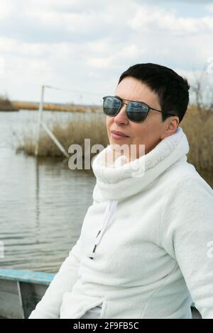 Una donna di mezza età gode dell'aria fresca all'inizio della primavera sul fiume. La donna indossa un pullover sportivo bianco. Foto Stock