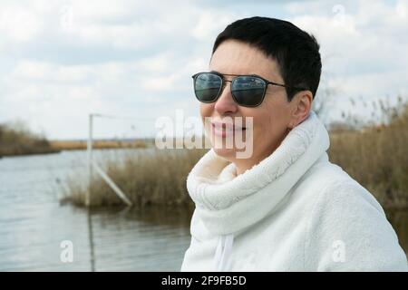 Una donna di mezza età gode dell'aria fresca all'inizio della primavera sul fiume. La donna indossa un pullover sportivo bianco. Foto Stock