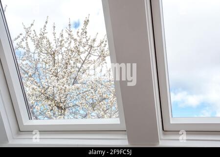 Un moderno lucernario aperto, finestra mansarda contro il cielo blu con un bell'albero con fiori bianchi, nuovo design moderno della casa, dettagli architettonici Foto Stock