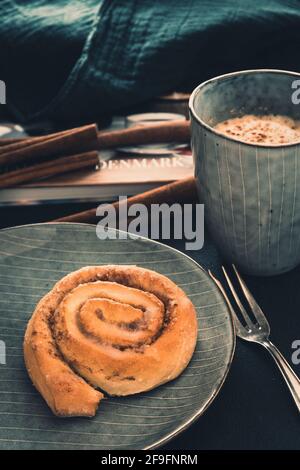 Rotolo di cannella su un piatto grigio, caffè e forchetta. Sullo sfondo un libro, bastoni di cannella e un panno, colori di moody. Primo piano verticale. Foto Stock