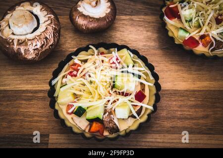 Primo piano di tartine di verdure crude con formaggio in piatti di crostata nera su un tavolo di legno, preparati per la cottura. Foto Stock