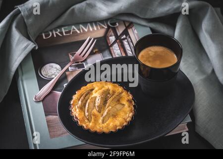 Tartlet di mela su un piatto nero e un caffè su un libro di tavolino preparato per una pausa. Colori pastello. Foto Stock