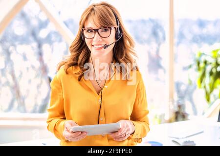 Donna d'affari matura in cuffia che parla con una chiamata in conferenza e tiene il tablet digitale in mano mentre lavora da casa. Ufficio domestico. Foto Stock