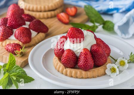 Piatto bianco con tartleton di fragole, decorato con fiori e foglie di menta, sullo sfondo tavola di legno con tartine e fragole Foto Stock