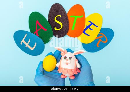 Tessera di Pasqua 2021. Uova di carta multicolore con l'iscrizione PASQUA e mani in guanti medici protettivi che tengono un uovo giallo e Pasqua fatti in casa Foto Stock