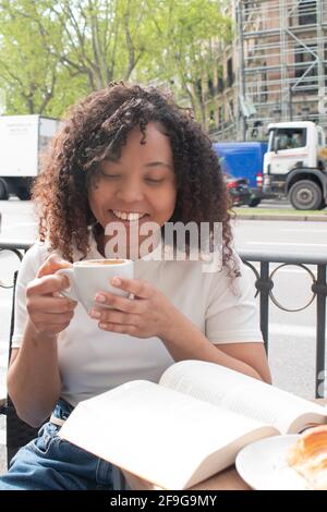 giovane donna nera con ricci che beve un caffè e che legge un libro all'esterno di una caffetteria. Foto Stock