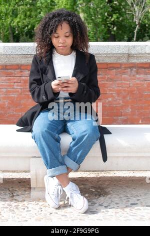 giovane donna nera con ricci seduti su una panca utilizzando il telefono cellulare all'esterno sullo sfondo di mattoni e alberi. Foto verticale. Foto Stock