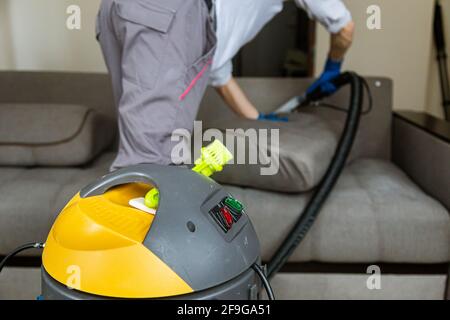 Servizio di pulizia. Uomo janitor in guanti e uniforme aspirapolvere divano con attrezzature professionali Foto Stock