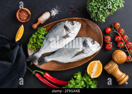 Pesce Dorado con verdure e spezie pronte per la cottura, vista da tavolo, fondo nero in cemento. Due pesci dorado crudi non cotti su vassoio di legno, Foto Stock