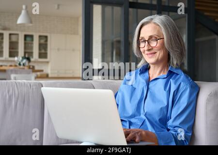 Donna anziana di 60 anni che usa tecnologie informatiche a casa seduta sul divano. Foto Stock
