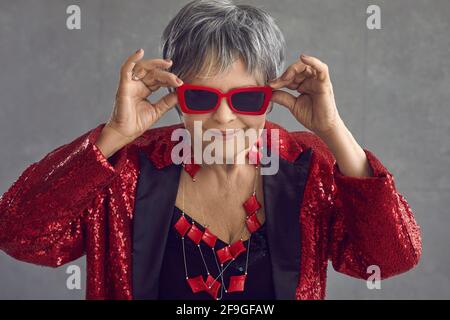 Ritratto di donna anziana divertente e fiduciosa in sequin rosso outfit e occhiali da sole freschi Foto Stock