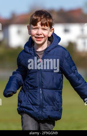 Un simpatico ragazzo rosso dagli occhi azzurri che indossa una giacca blu che corre in un parco in una soleggiata giornata primaverile Foto Stock