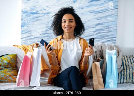 Eccitata gioiosa ragazza curly afroamericana graziosa, felice con nuovi acquisti, seduta sul divano a casa, utilizza smartphone e carta di credito per il pagamento online, vicino a un sacco di borse di carta, sorridere felicemente Foto Stock