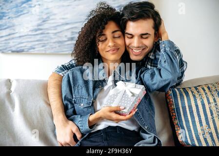 Regalo di compleanno o anniversario. Ragazzo di razza mista dà una scatola con un regalo alla sua amata bella ragazza afroamericana, fa una sorpresa, abbracciandosi l'un l'altro su un divano a casa, felice rapporto Foto Stock