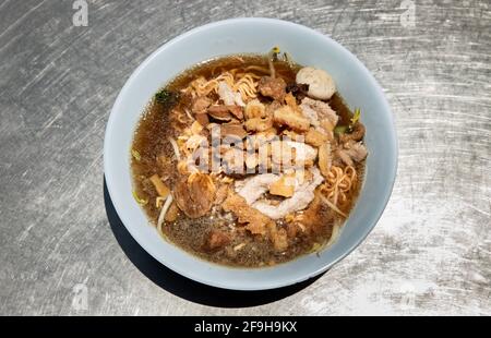 Spaghetti istantanei in zuppa di maiale brasata con palle di maiale e scorze di maiale grasso in ciotola bianca. Cucina tailandese, vista dall'alto, fuoco selettivo. Foto Stock