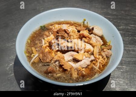 Spaghetti istantanei in zuppa di maiale brasata con palle di maiale e scorze di maiale grasso in ciotola bianca. Cibo tailandese, fuoco selettivo. Foto Stock