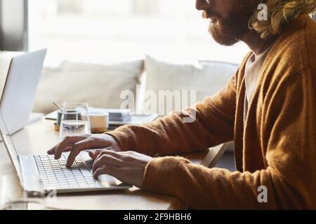 Minimo ritaglio di uomo bearded utilizzando il computer portatile mentre si lavora al bar tavolo illuminato dalla luce del sole in calde tonalità marrone, spazio di copia Foto Stock