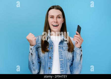 Ecited giovane donna tiene smartphone ricevere sms con grandi notizie si sente felice fare sì gesto, indossa una giacca denim, isolato su sfondo blu Foto Stock