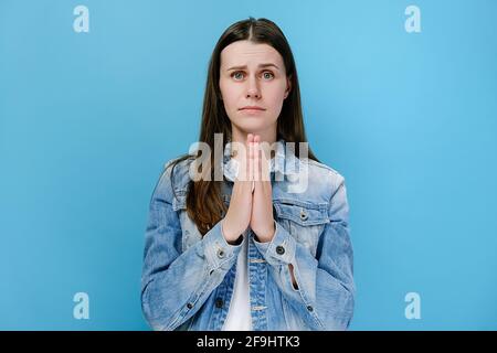 Ritratto di giovane donna che chiede di tenere le mani piegate gesto di preghiera che implora qualcosa dire per favore, vestito in giacca denim, isolato su parete blu Foto Stock