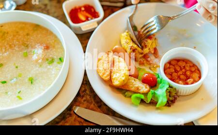 La persona usa forchetta e cucchiaio per la colazione al ristorante. Porridge di riso o gruel di riso con gamberi in ciotola bianca e fette di salsiccia e patate fritte. Foto Stock