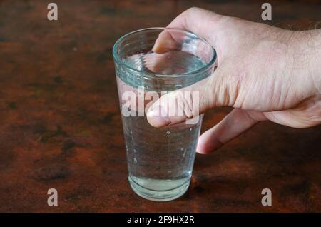 Un bicchiere con acqua e una mano dell'uomo su un tavolo rosso. Un vetro trasparente pieno d'acqua e la mano destra di un maschio caucasico adulto. All'interno. Lifestyl Foto Stock