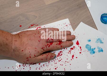 La mano sinistra di un maschio adulto schizzato con vernice rossa sulla parte superiore. L'uomo mise la sua palma su un foglio bianco di carta sul pavimento. Un footprint blu per animali domestici Foto Stock