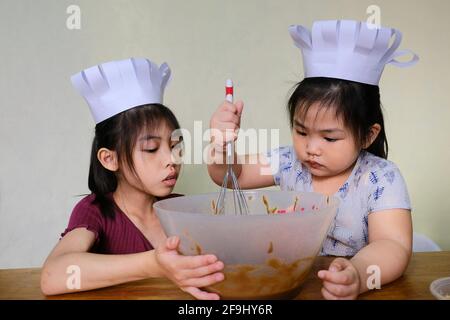 Due carine giovani sorelle asiatiche con simpatici cappelli da chef bianchi, stanno imparando a preparare i biscotti, aiutandosi a vicenda mescolando l'impasto in una grande ciotola con una mano Foto Stock