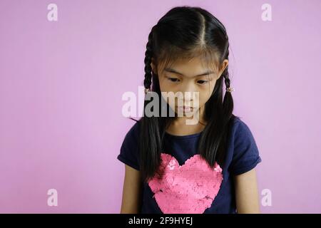 Un colpo di mezzo corpo di una ragazza asiatica carina giovane che guarda giù, sentendosi sconvolto, contemplando la sua azione seguente. Sfondo rosa brillante. Foto Stock