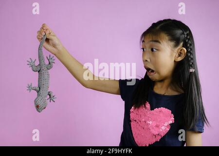 Una giovane ragazza asiatica carina è stata pranked con un gecko realistico, tenendolo dalla coda, comportandosi sorpreso e spaventato. Foto Stock