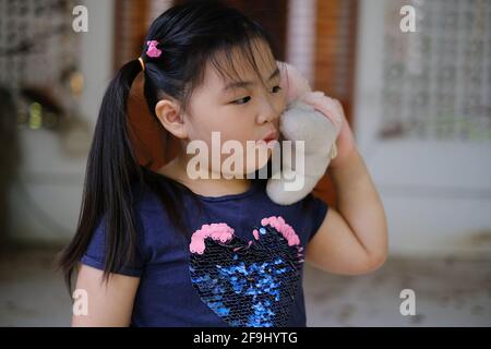 Una giovane ragazza asiatica carina sta tenendo il suo orsacchiotto bianco, fingendo che si tratta di un telefono cellulare e giocosamente parlando nella bambola, divertendosi, sorridendo Foto Stock