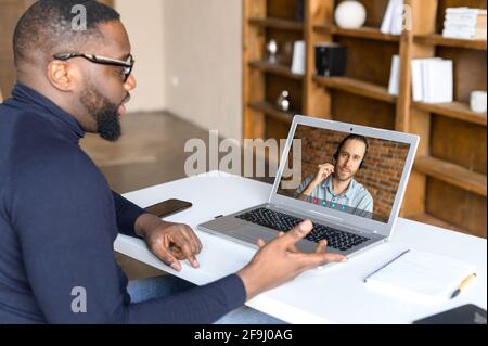 Uomo d'affari afro-americano si sente felice di aver chiamato con un collega maschio in cuffia utilizzando un'applicazione speciale, parlare di fronte alla webcam, briefing online o riunione, impegnato in conferenza web con il partner Foto Stock