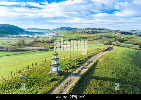 Fuco di un monumento cristiano e percorso che porta a una piccola città con vaste montagne verdi e nebbie sullo sfondo. Foto Stock