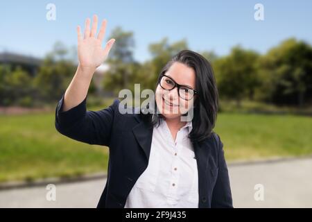 Donna d'affari adulta allegra che indossa occhiali o occhiali e casual elegante tuta da ufficio che svela la mano mentre saluta il gesto nel parco natura backgroun Foto Stock